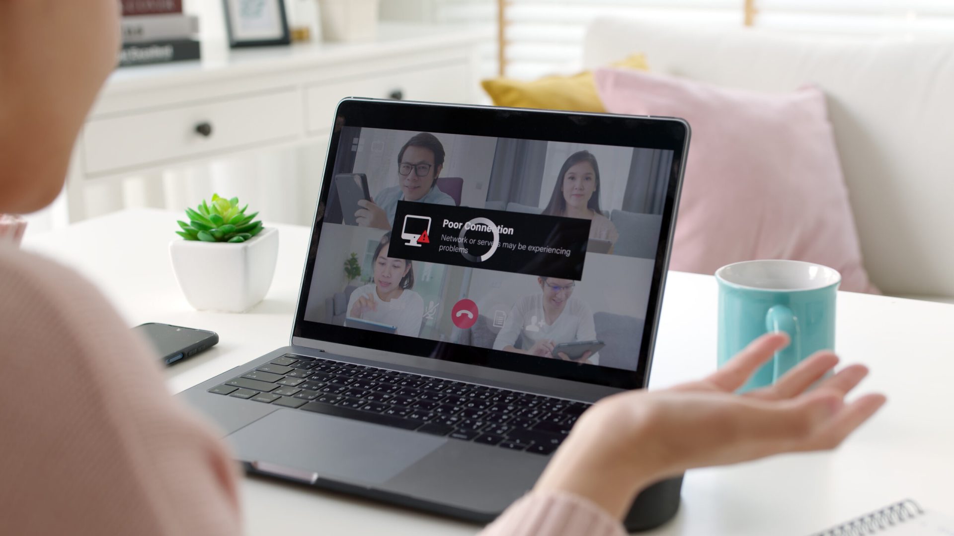 A person views a laptop screen displaying a video call with multiple participants, showing a "Poor Network Connection" warning. The setting is a cozy home office with a succulent plant, a mug, and a notebook on the desk