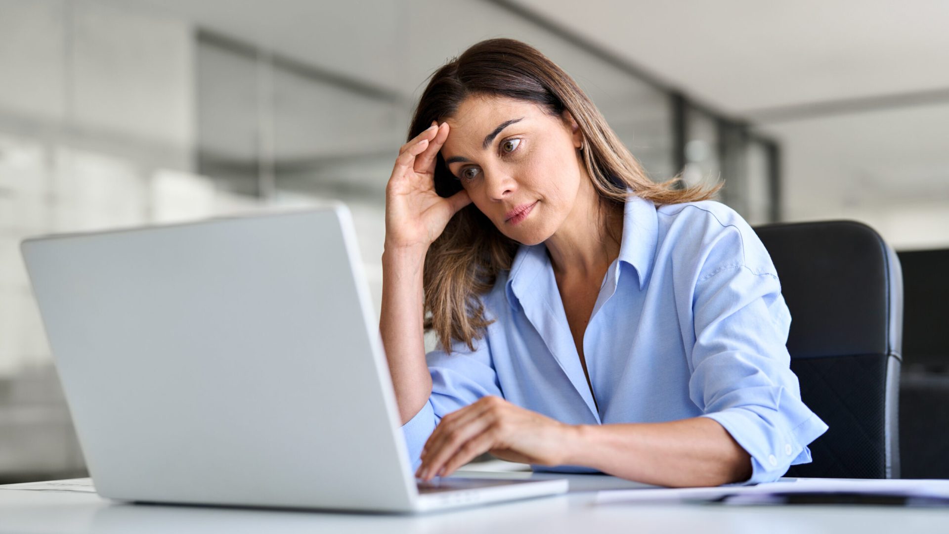 A woman in a blue shirt sits at a desk in a modern office, resting her head on her hand with a thoughtful expression while working on a laptop. The background is softly blurred, suggesting a quiet workspace